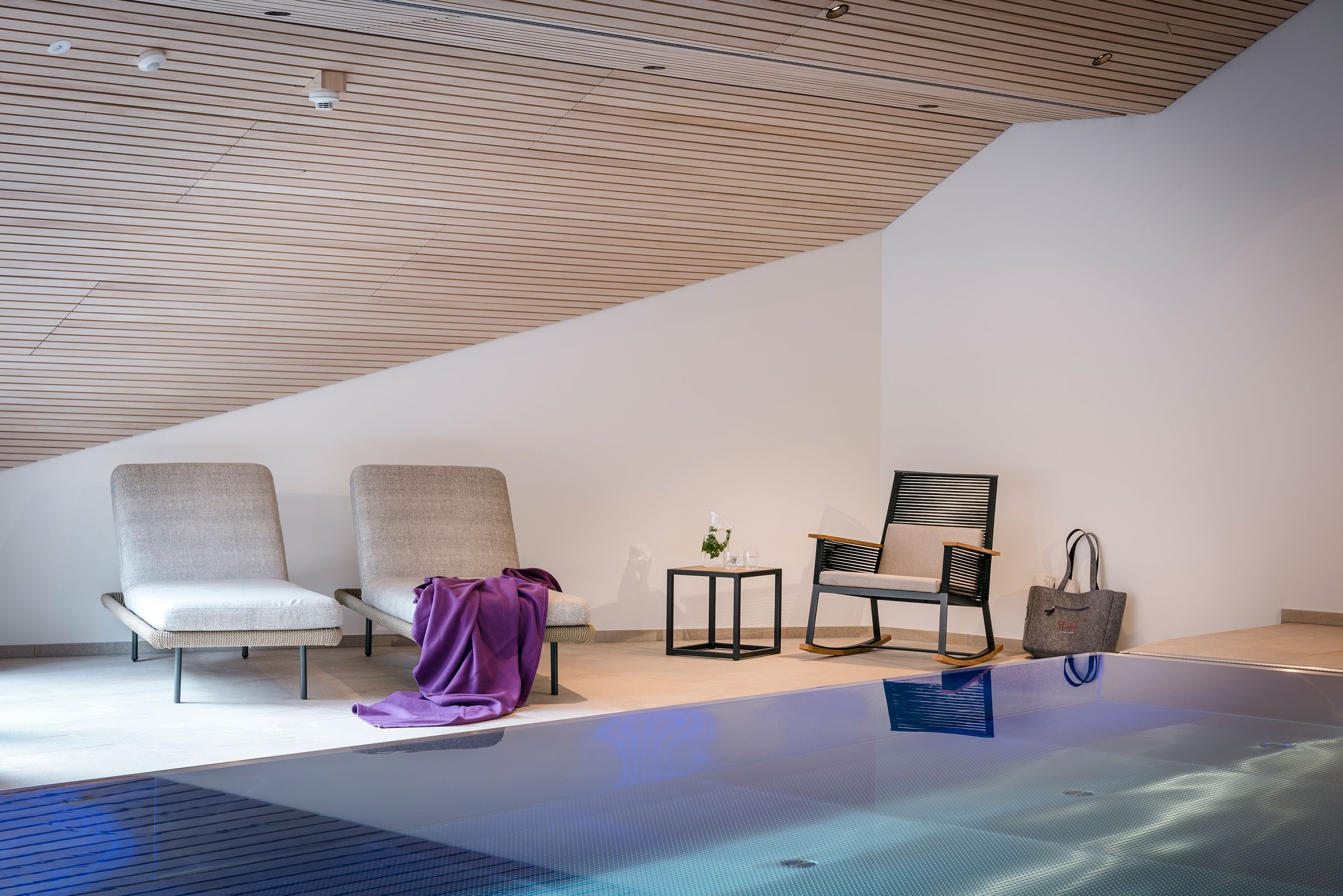 Un espace piscine intérieur moderne, comprenant deux chaises longues, une petite table et une chaise avec un sac à main.