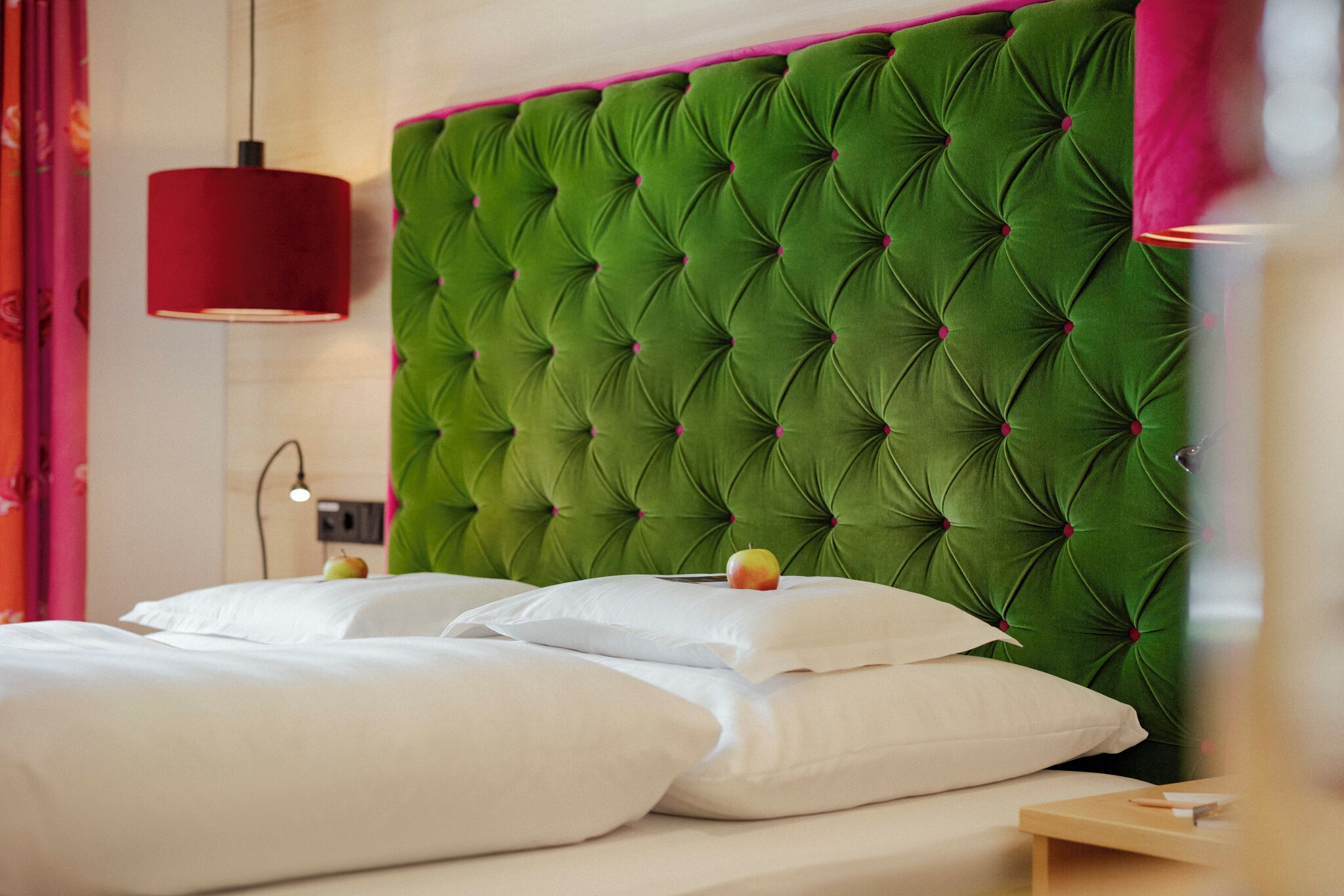 A hotel room with a green tufted headboard, white bedding, and a red pendant light.