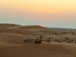 Paisaje desértico con dunas de arena y un atardecer. Varias tiendas de campaña están instaladas con sillas y mesas para acampar.