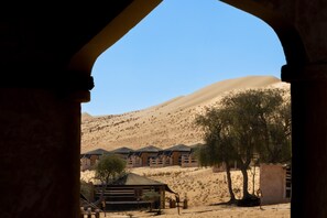 A desert landscape with a building featuring multiple rooms with sloped roofs, surrounded by sand dunes and sparse vegetation. - Thousand Nights Camp (Bidiya)