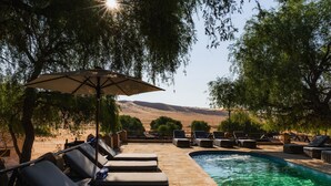 Zona de piscina amb gandules i un parasol, envoltada d'arbres i vista a les muntanyes.