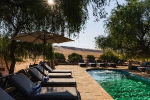 A poolside area with lounge chairs and an umbrella, surrounded by trees and a view of the hills.