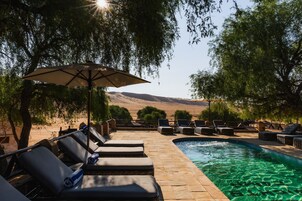 Zona junto a la piscina con sillones de descanso y una sombrilla, rodeada de árboles y vistas a las colinas.