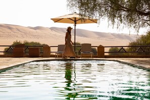 Zona de piscina con sillas de descanso y una sombrilla, rodeada de colinas desérticas.