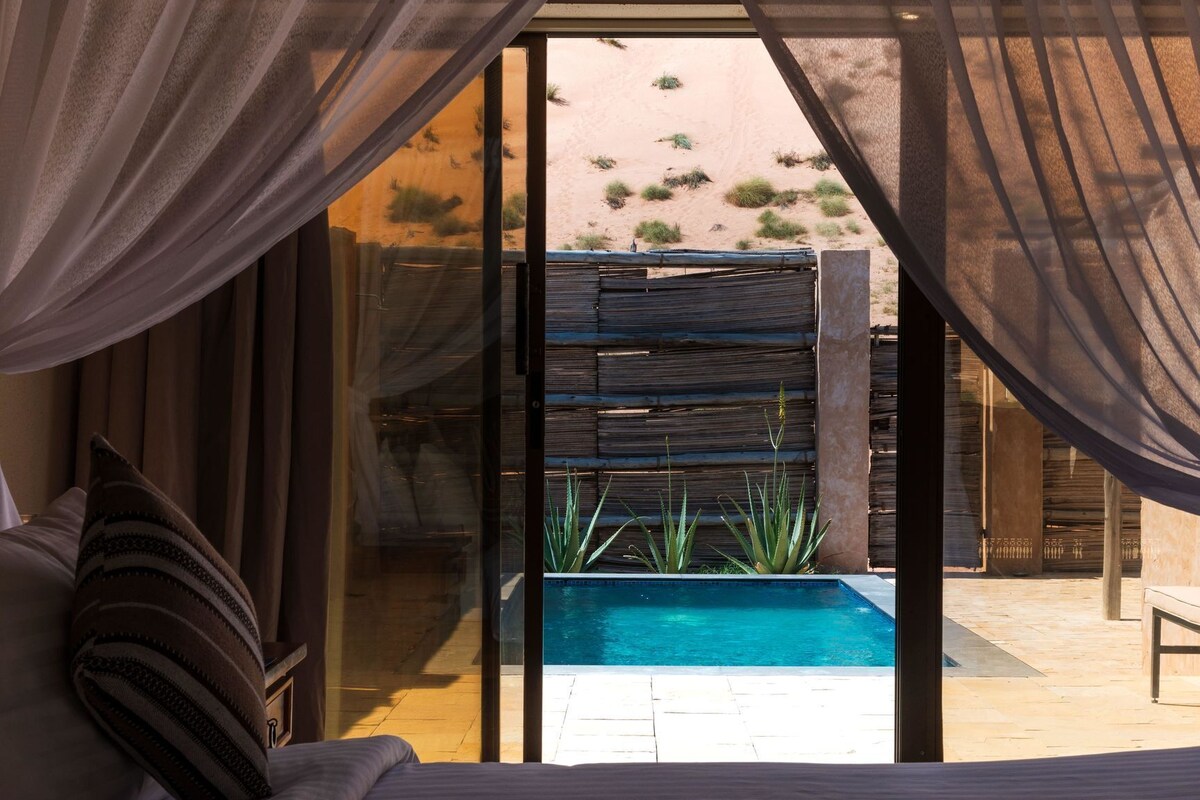 A bedroom with a large bed, a view of a swimming pool, and a wooden fence outside.