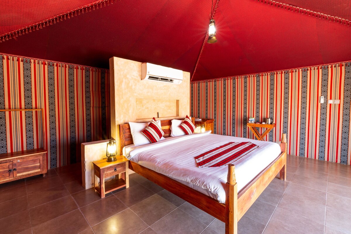 A spacious bedroom with a large bed, a wooden headboard, a nightstand, a lamp, and a wall-mounted air conditioner.
