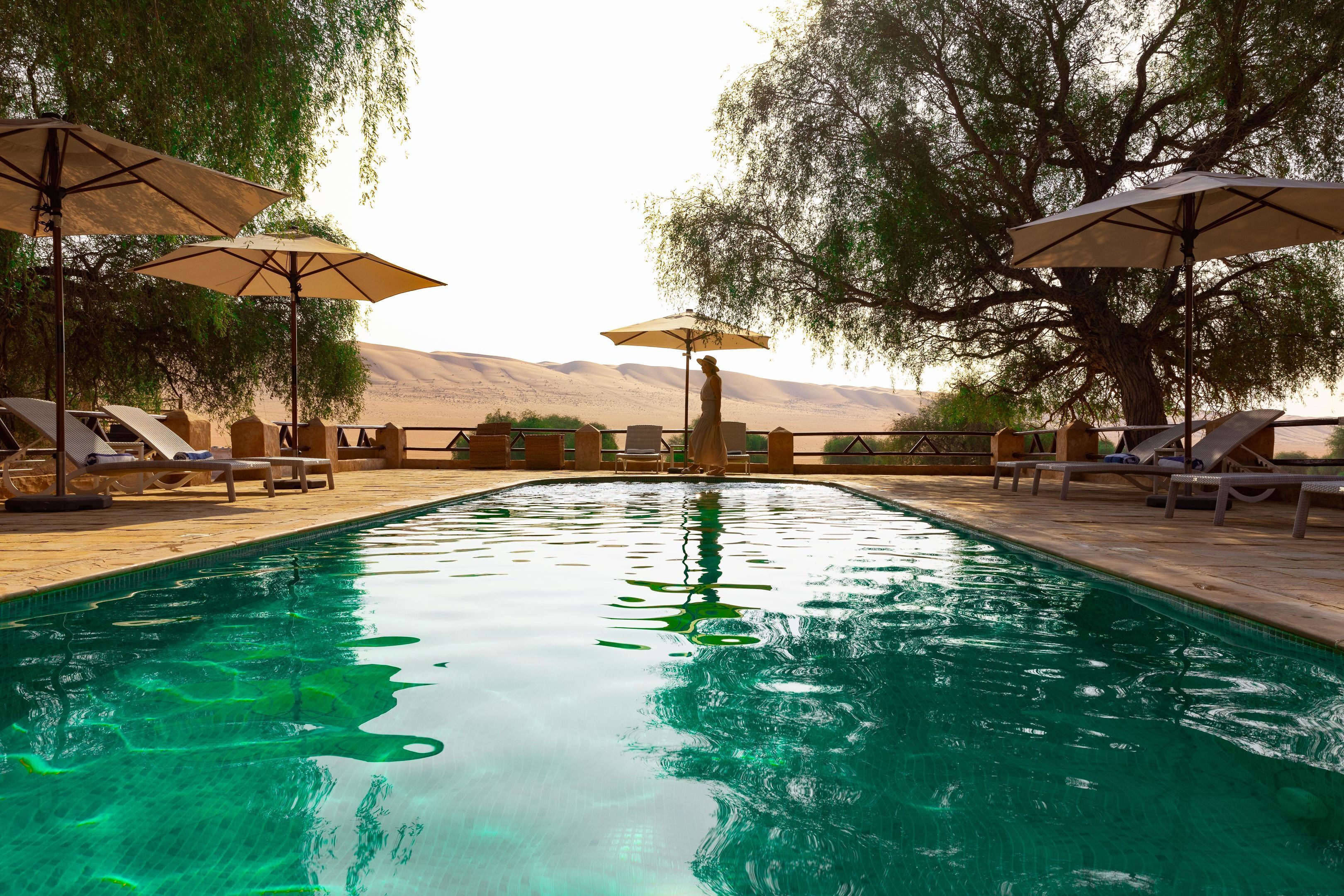Une piscine entourée de chaises longues et de parasols, bordée d’arbres et avec un paysage désertique en arrière-plan.