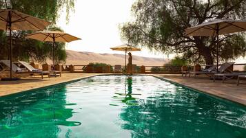 Une piscine entourée de chaises longues et de parasols, bordée d’arbres et avec un paysage désertique en arrière-plan.