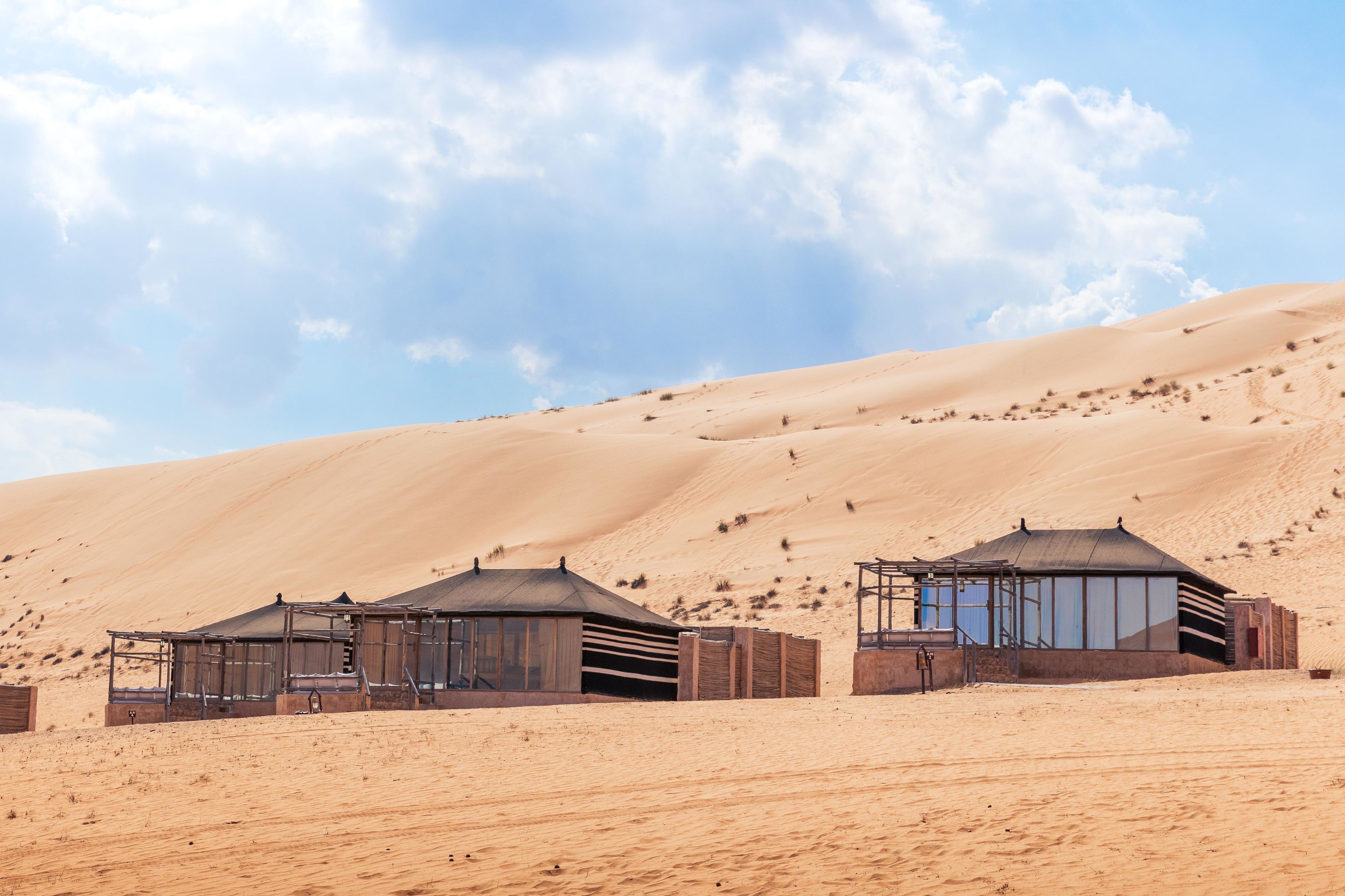Wüstenlandschaft mit zwei modernen, minimalistischen Gebäuden, die große Fenster und flache Dächer haben, umgeben von Sanddünen.