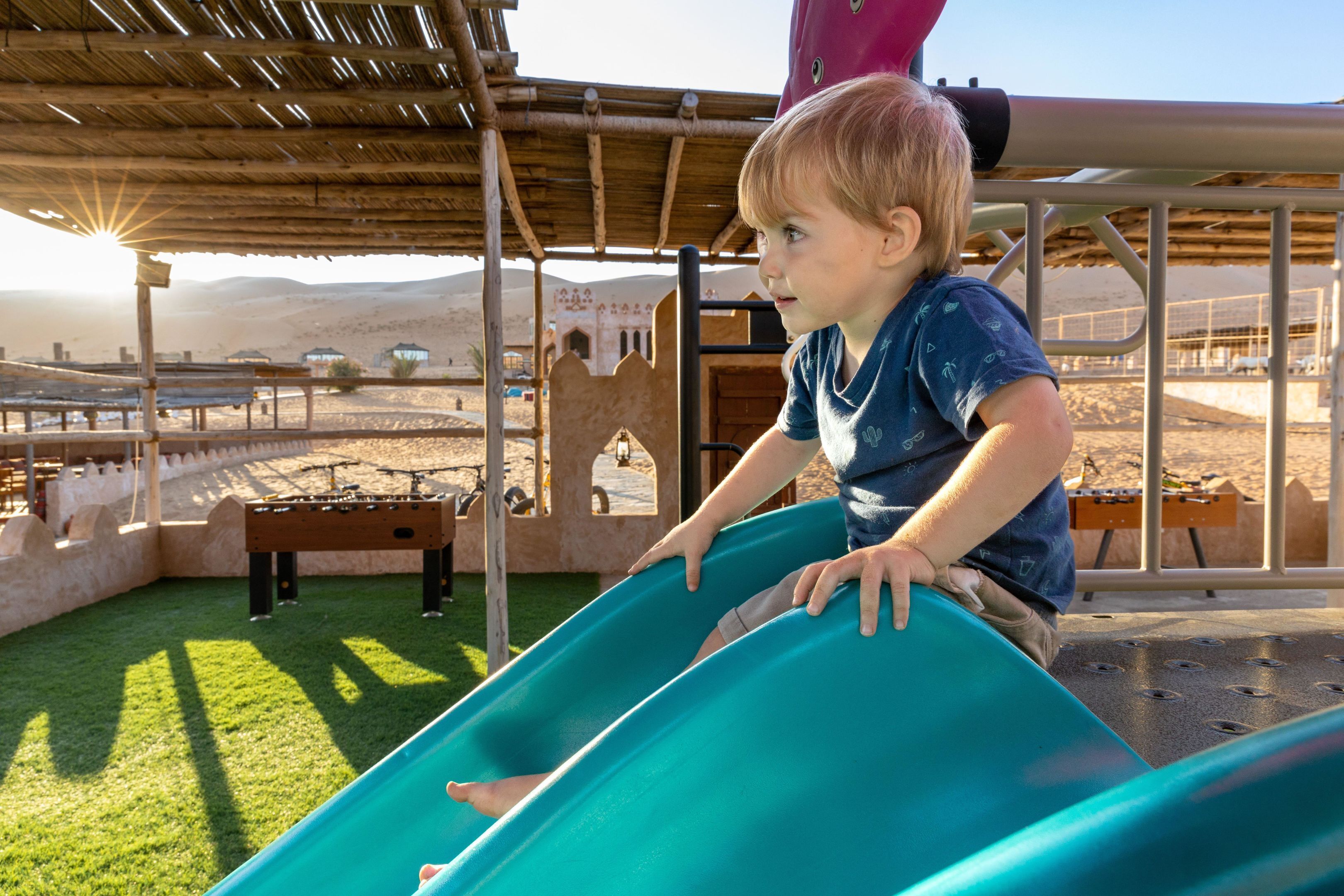 A child on a slide in an outdoor play area with a view of a desert landscape.