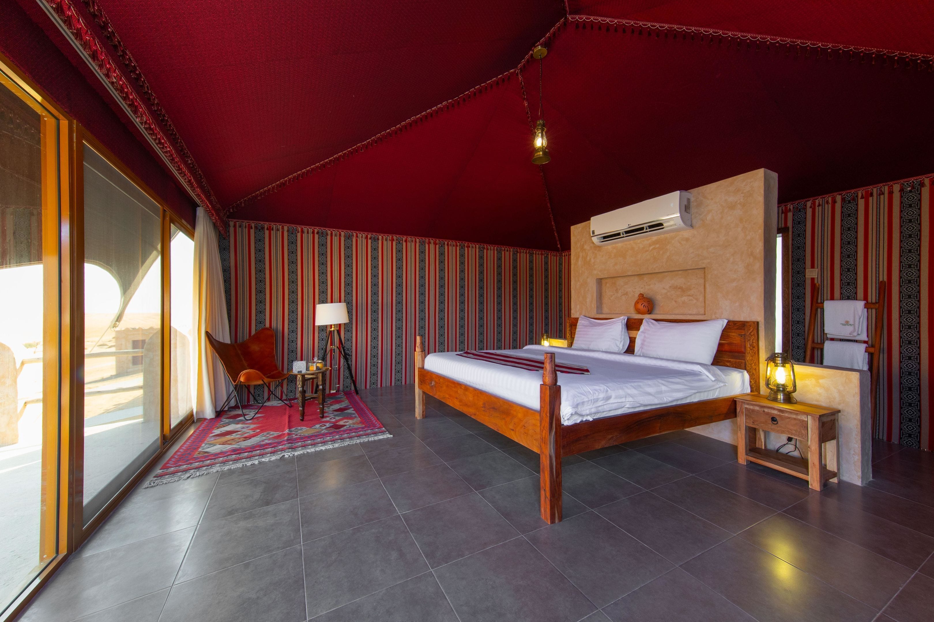 A spacious bedroom with a large bed, a nightstand, a lamp, and a view of the outdoors through large windows.
