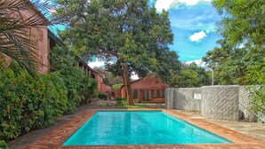 Una piscina rectangular con borde de ladrillo, rodeada de exuberante vegetación y un edificio de ladrillo.