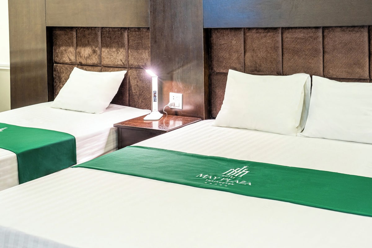 A hotel room with two beds, each with a green bedspread and white pillows. There is a nightstand with a lamp between the beds.