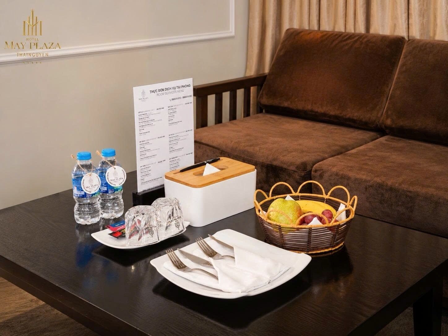 Habitación de hotel con un sofá marrón, una mesa de madera con una cesta de fruta, botellas de agua, una caja de pañuelos y un cartel con el menú.