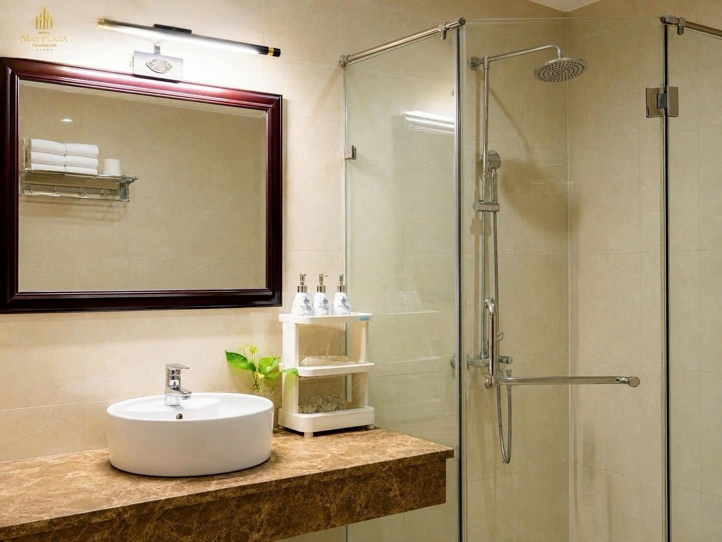 Un baño de hotel con ducha de mampara de vidrio, un lavabo redondo, un espejo con marco de madera y un estante con artículos de aseo.