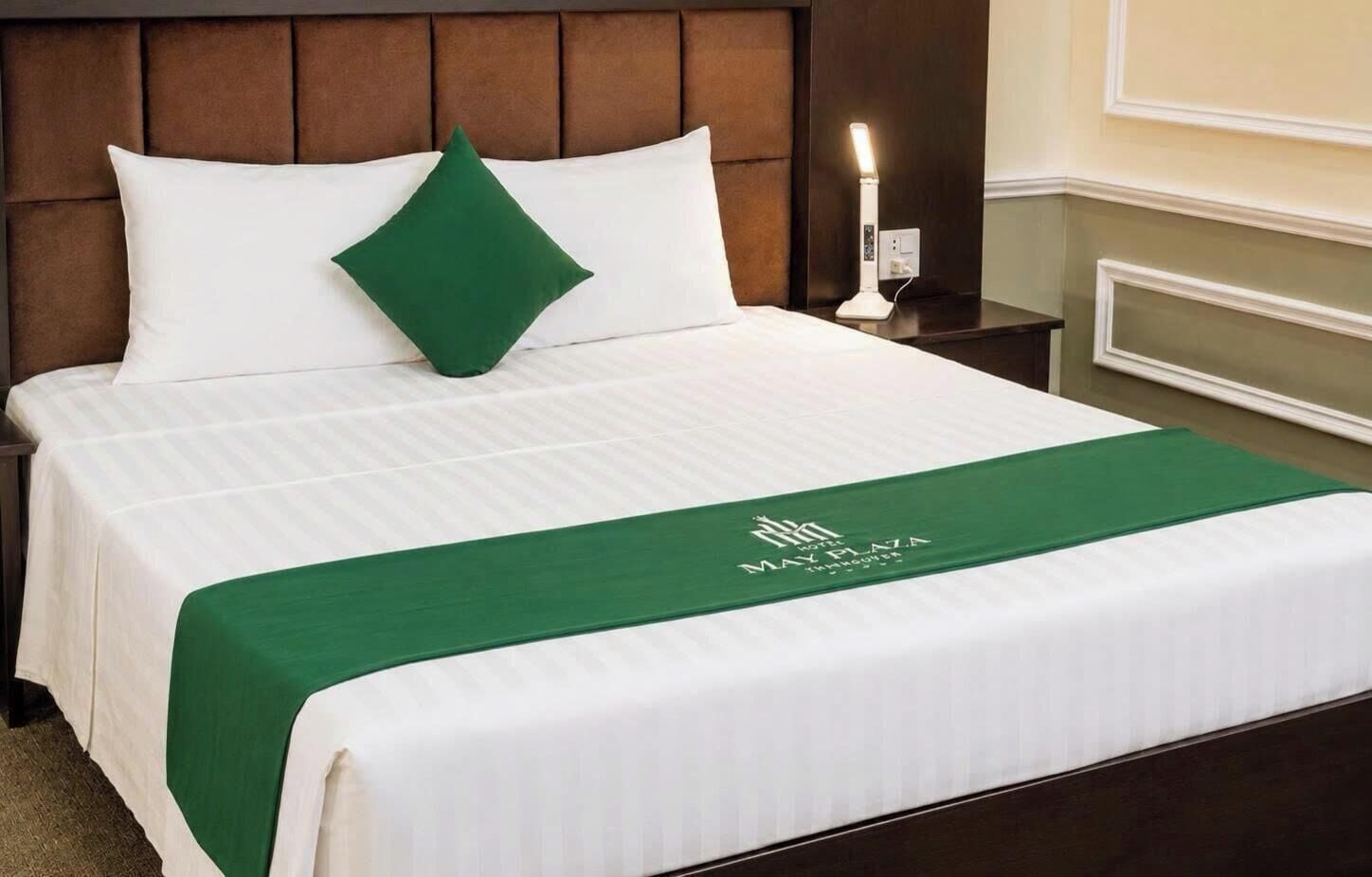 Un lit bien fait, avec une couverture verte et un oreiller, une table de chevet avec une lampe, et un mur orné d’un panneau décoratif.