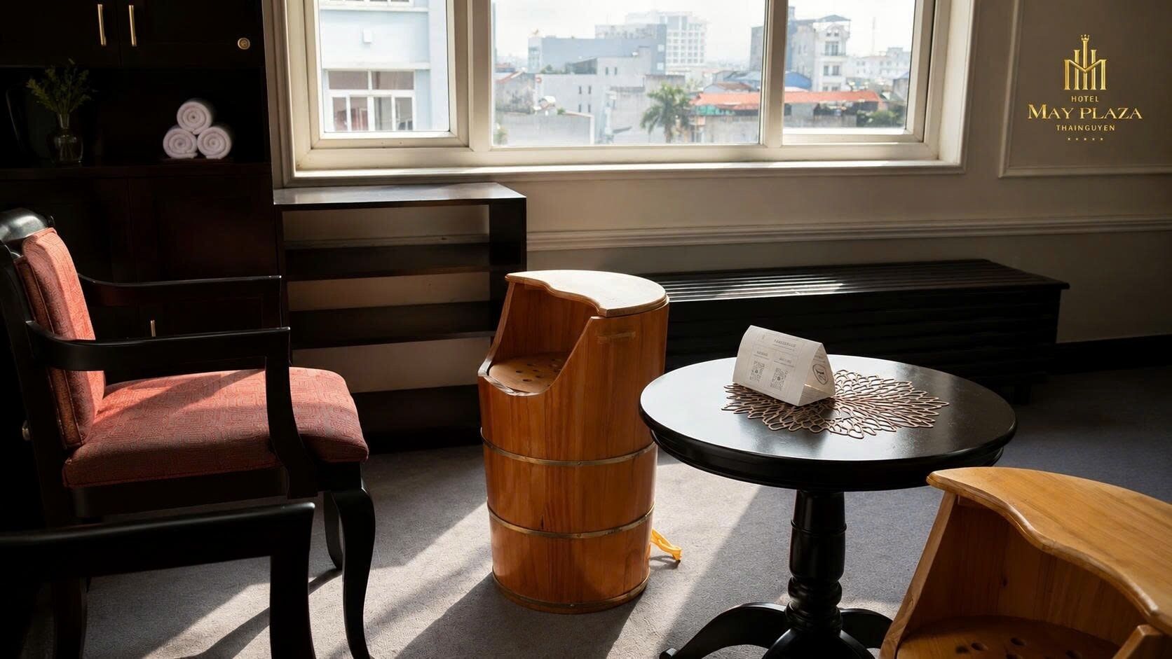 Quarto de hotel com vista para a cidade, um banquinho de madeira, uma mesa redonda com jogo americano e uma cadeira estofada vermelha.