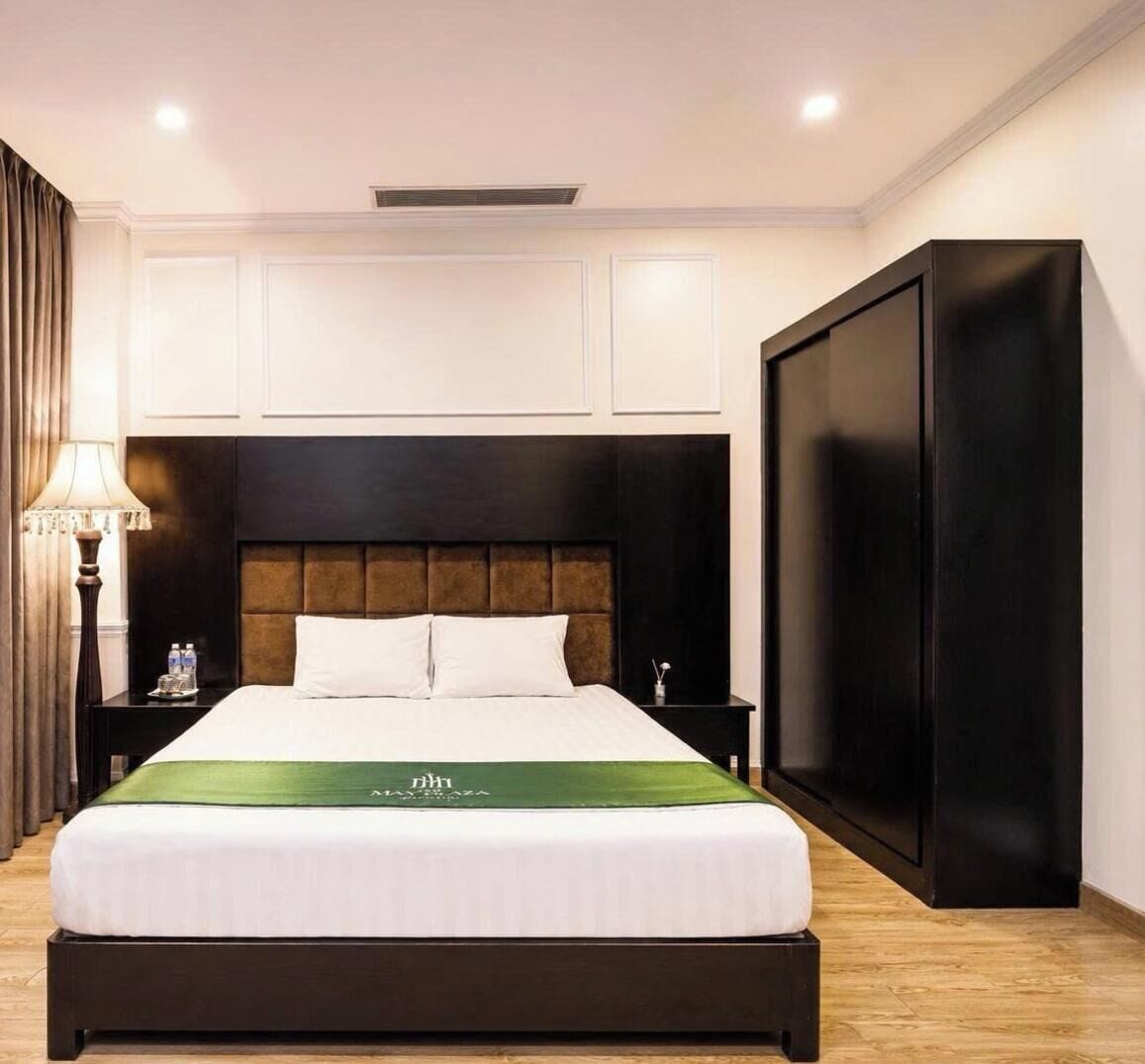 A hotel room with a large bed, a nightstand with a lamp, a wardrobe, and a wall-mounted air conditioning unit.