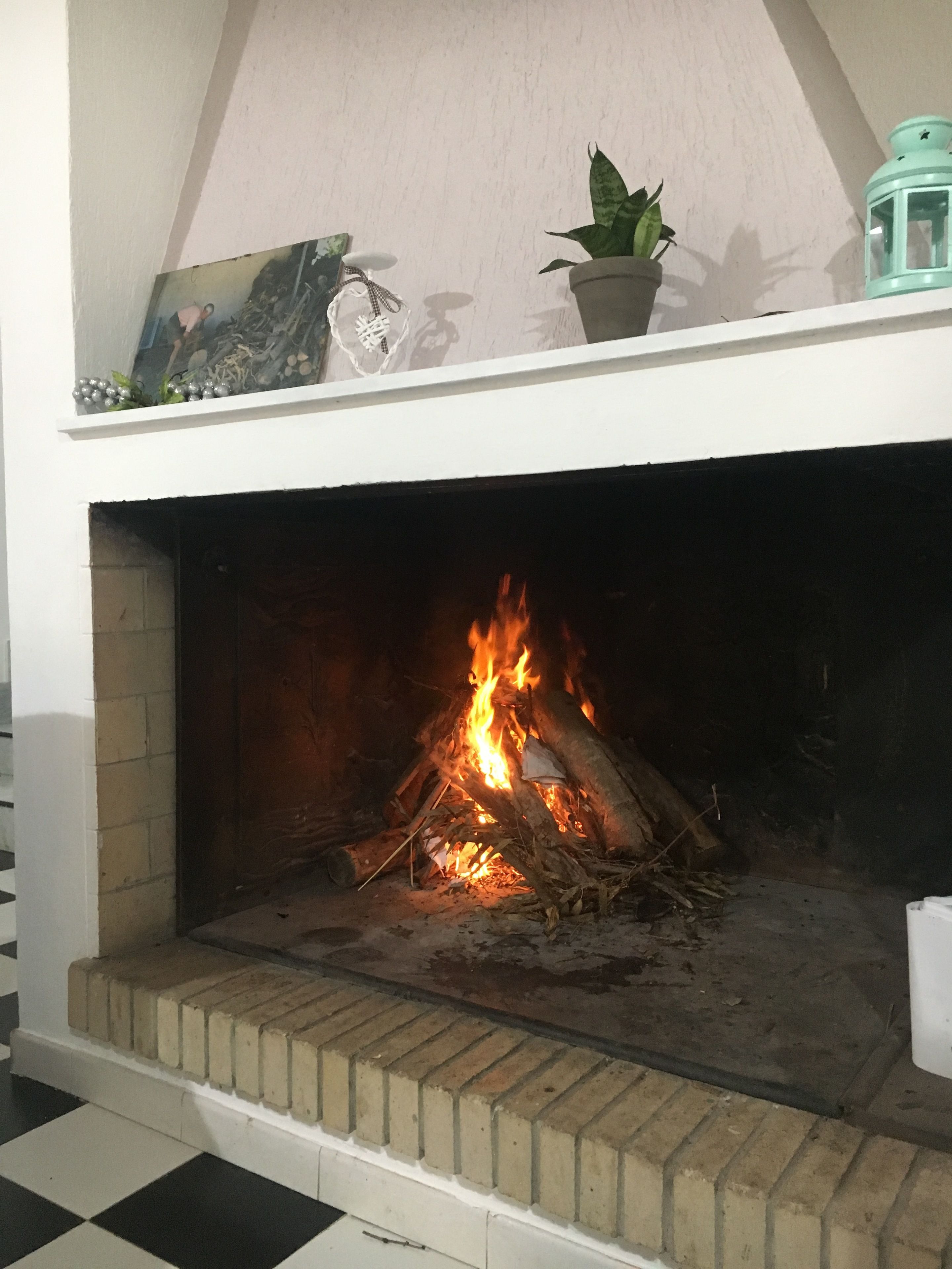 Una chimenea con fuego encendido, rodeada por una repisa blanca con una planta en maceta y un farol.