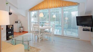 A modern living room with a glass coffee table, white furniture, a flat-screen TV, and large windows with blinds.