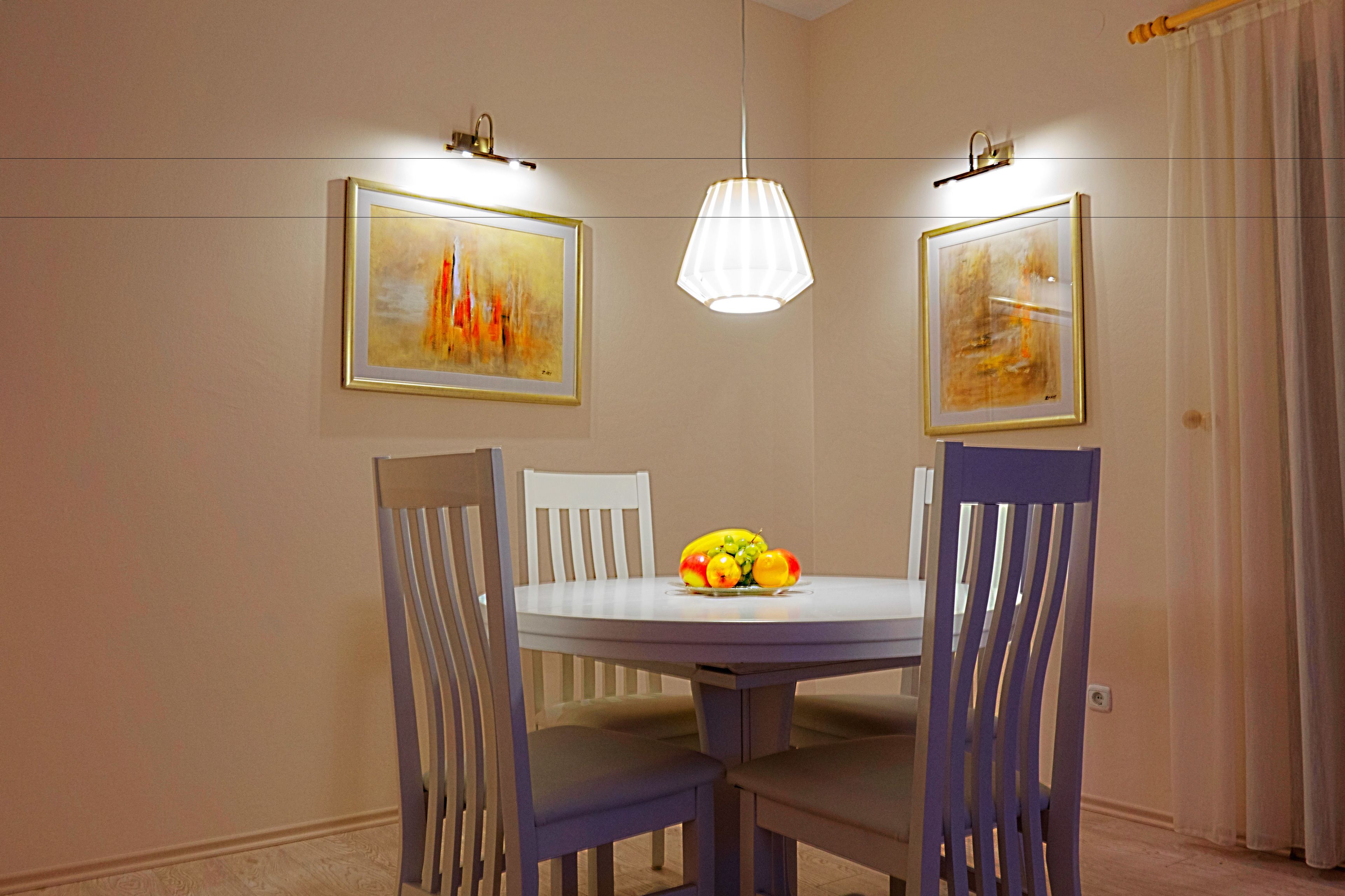 A dining area with a round table, four chairs, two framed paintings, and a pendant light.