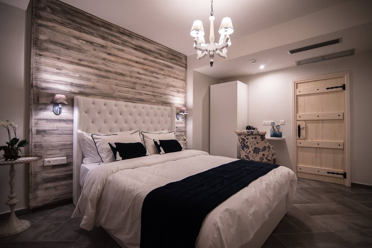 A bedroom with a tufted headboard, a chandelier, a bedside table with a lamp, and a wooden wardrobe.