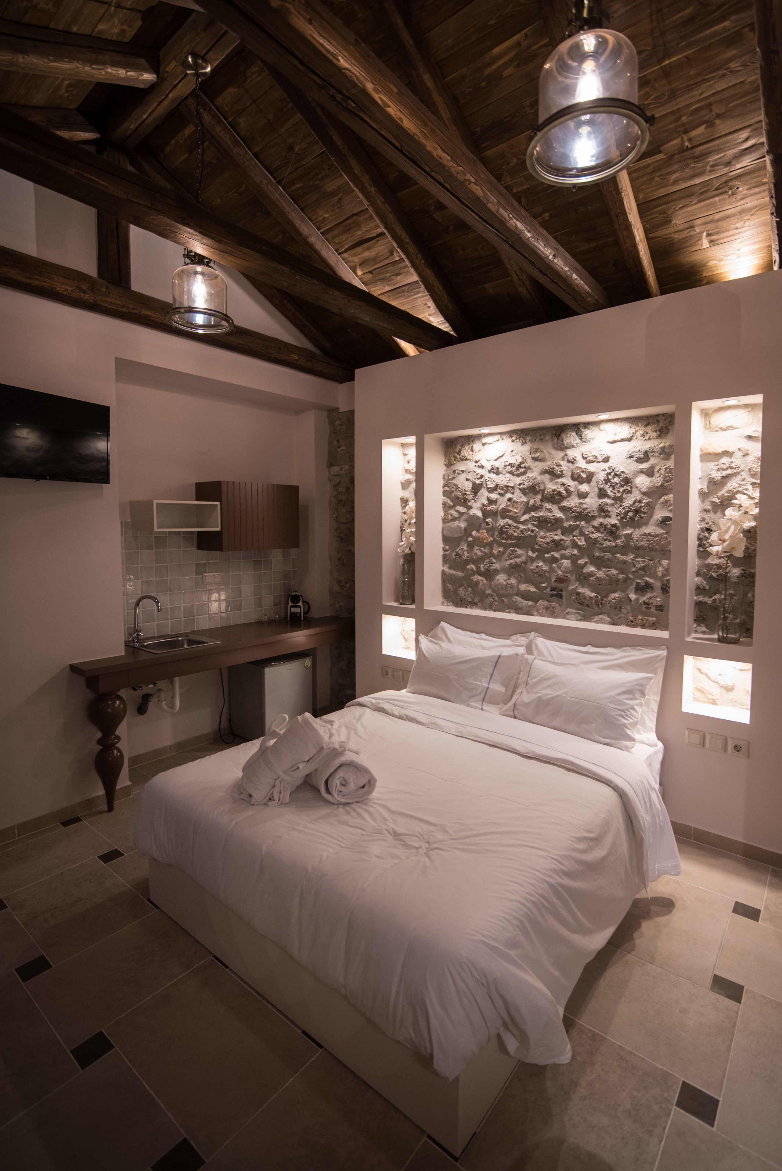 A bedroom with a large bed, a small kitchenette, and a stone wall with windows.