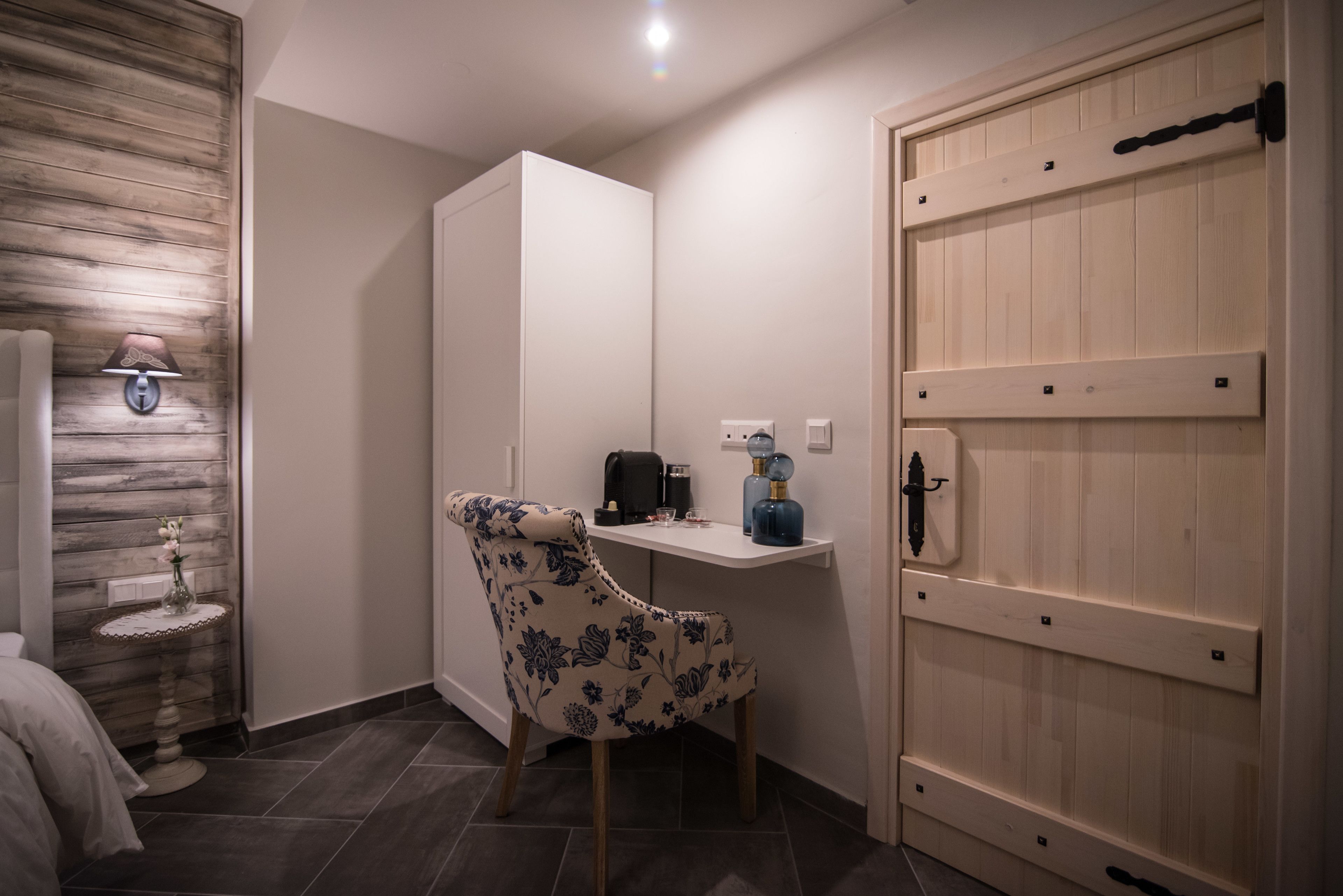 Una habitación pequeña y ordenada, con un muro de madera, un armario blanco, un sillón con estampado floral, una mesita con una lámpara y una puerta de madera.