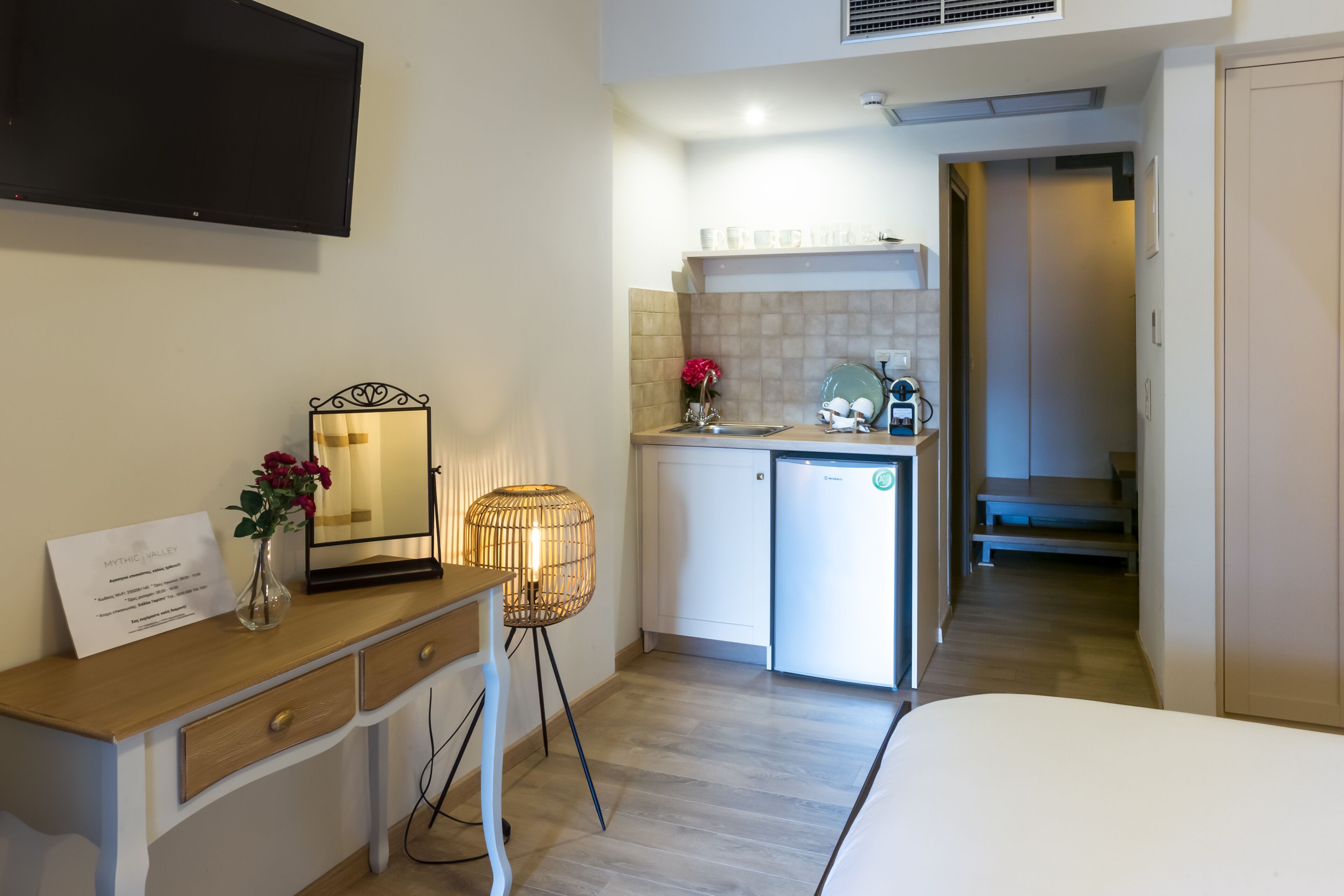 A compact hotel room with a bed, a small desk with a lamp, a bedside table with a vase of flowers, a compact kitchenette, and a bathroom visible through an open door.