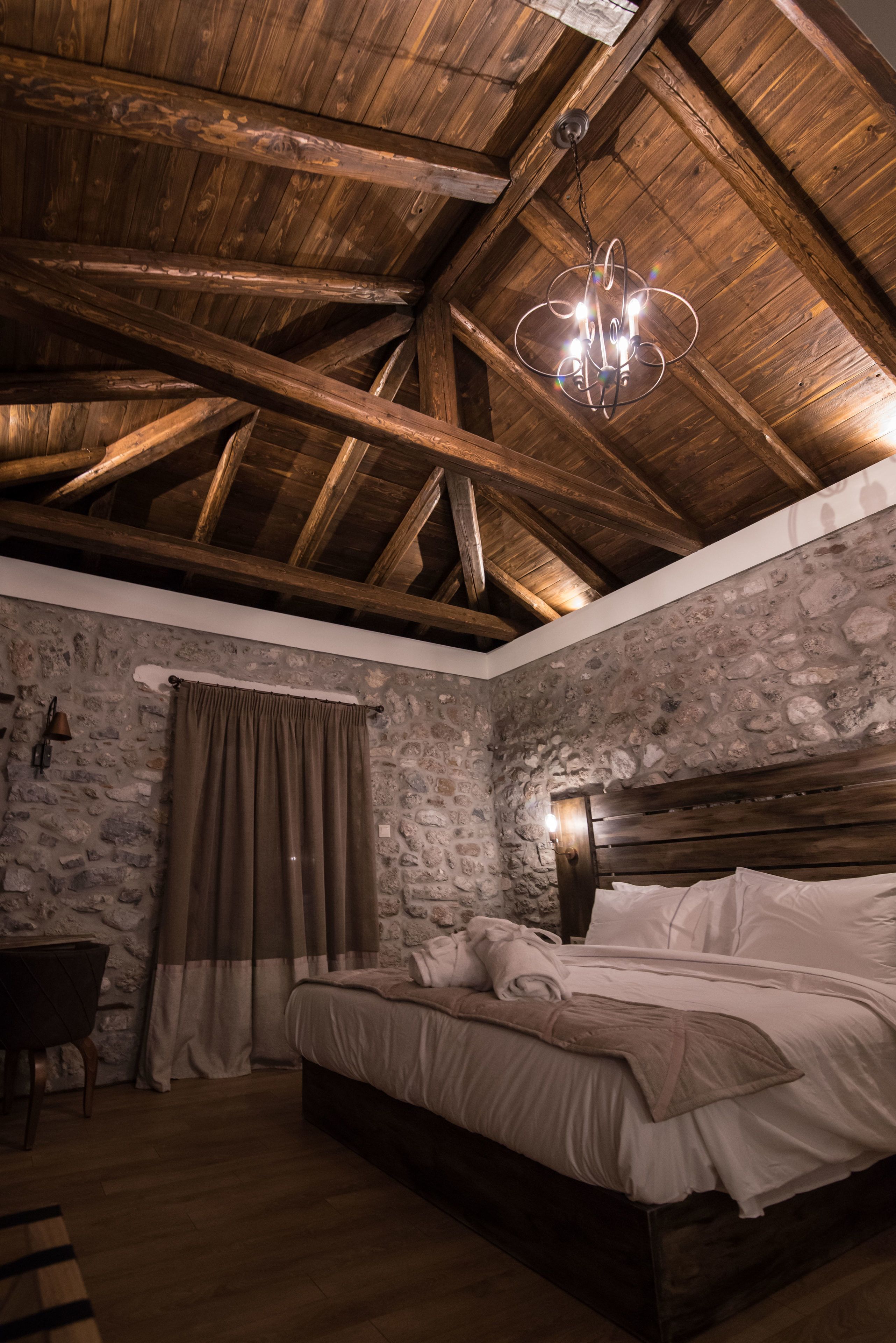 A bedroom with a stone wall, wooden ceiling beams, a bed, and a hanging light fixture.