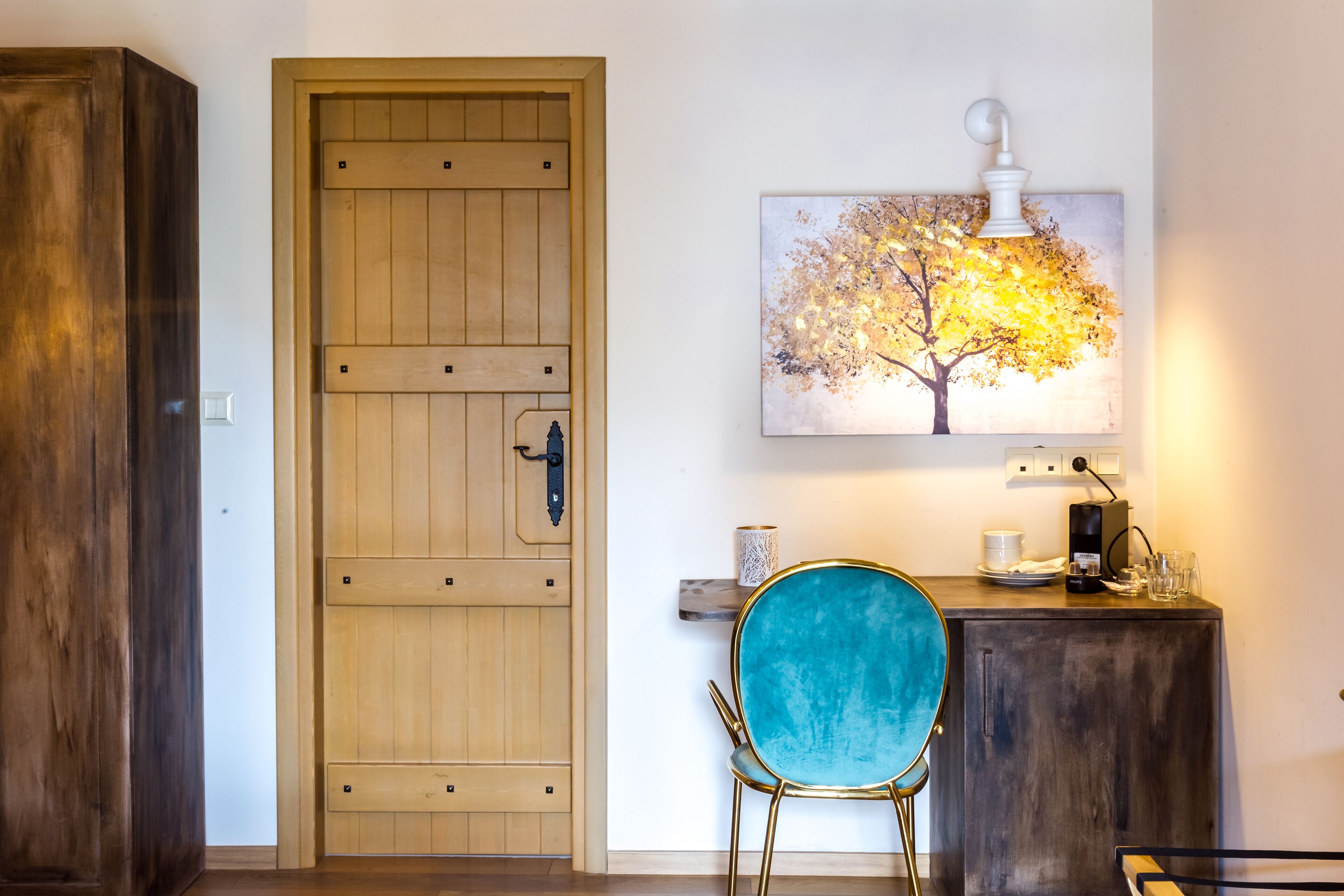 Una puerta de madera, una silla azul y un aparador con una lámpara y un cuadro de un árbol.