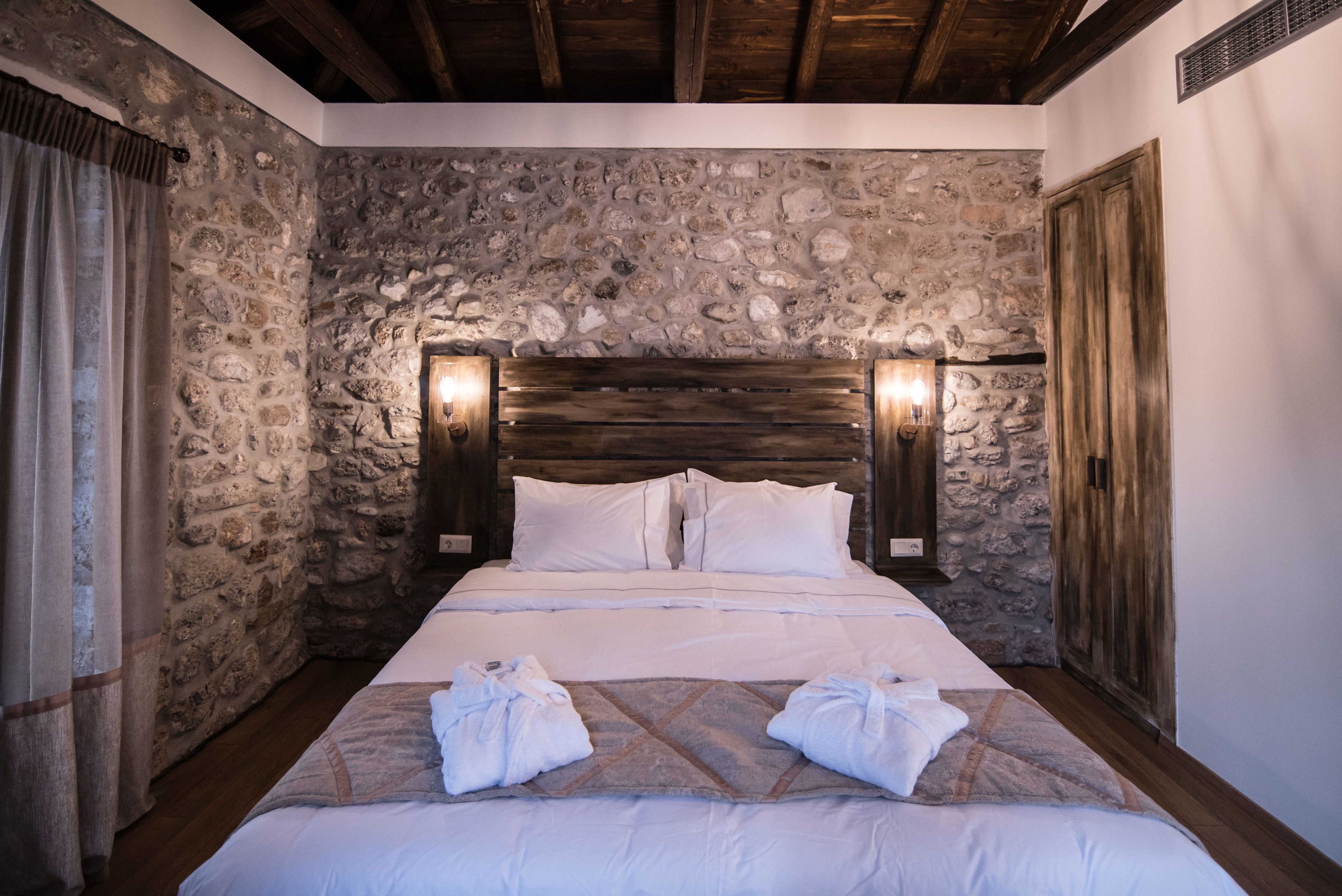 Una habitación con pared de piedra, vigas de madera en el techo, una cama con ropa blanca y dos lámparas de pared.