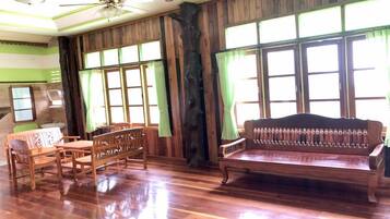 A room with wooden flooring, a ceiling fan, a wooden bench, a table with chairs, and large windows with curtains.