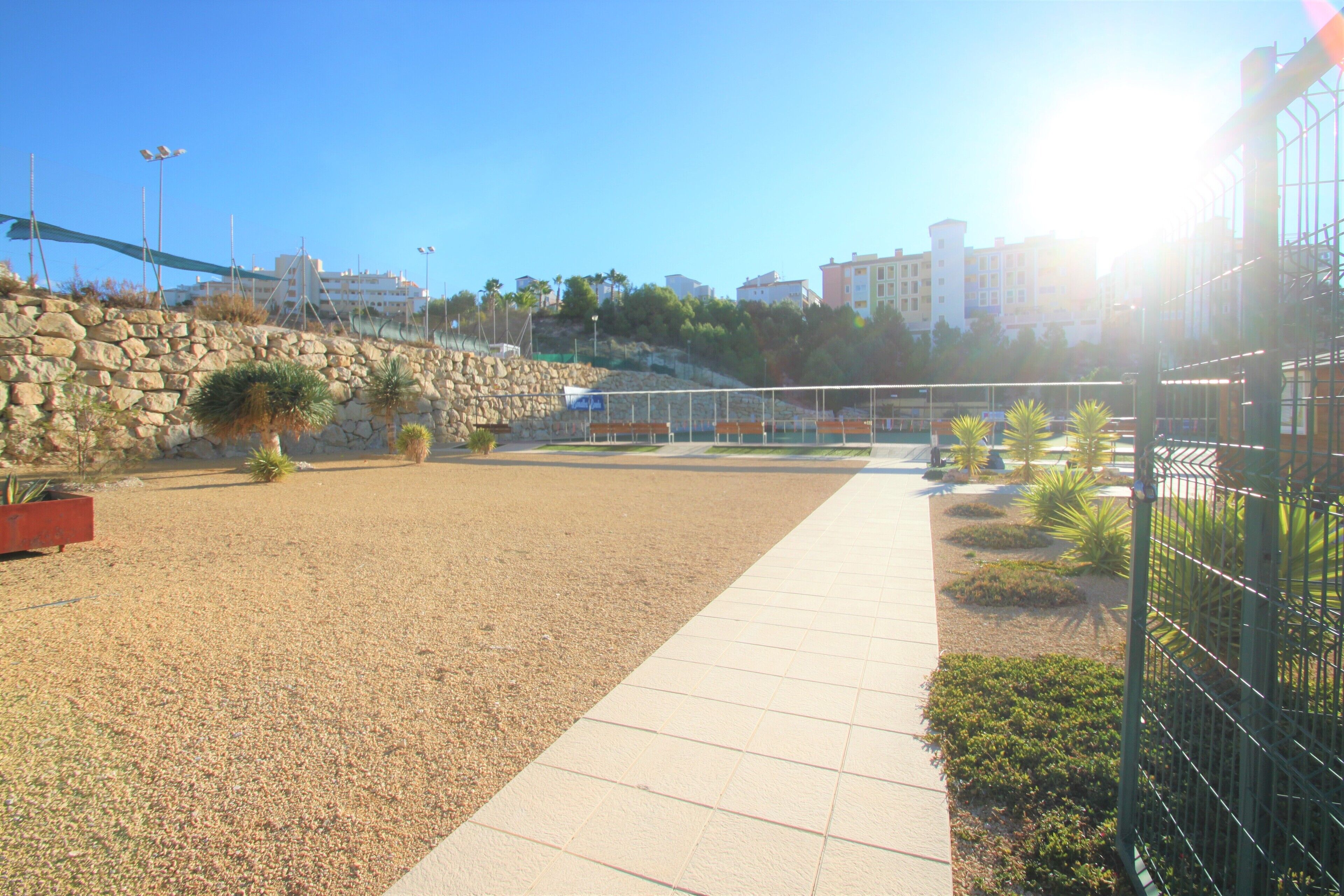 A paved walkway beside a sandy area with a stone wall and some greenery.