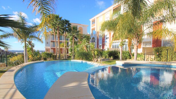 A swimming pool with a curved edge, surrounded by palm trees and a building with balconies. - Apartamento CasaTuris Bonalba Golf (Mutxamel)