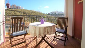 A balcony with a table and two chairs, a purple watering can, and a view of buildings on a hill. - Apartamento CasaTuris Bonalba Golf (Mutxamel)
