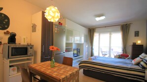 A compact hotel room with a kitchenette, a bed with striped bedding, a dining table with a vase of flowers, and a view of the outside through a window. - Apartamento CasaTuris Bonalba Golf (Mutxamel)