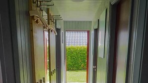 A corridor with a tiled floor leading to a door with a view of a green hedge and a tiled roof. - Juwel Feriendorf - Bockholm Haus 19 (Gluecksburg)