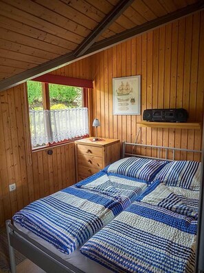 Ein Zimmer mit Holzwänden, einem Bett, einem kleinen Tisch, einer Lampe und einem gerahmten Bild an der Wand.