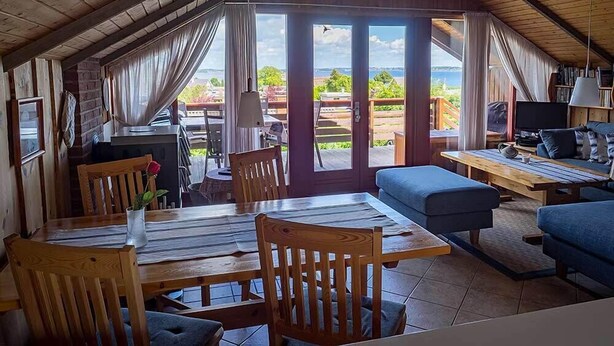 A cozy, open-plan living area with a dining table, chairs, a sofa, and a TV. Large windows offer a view of the outdoors. - Juwel Feriendorf - Bockholm Haus 19 (Gluecksburg)