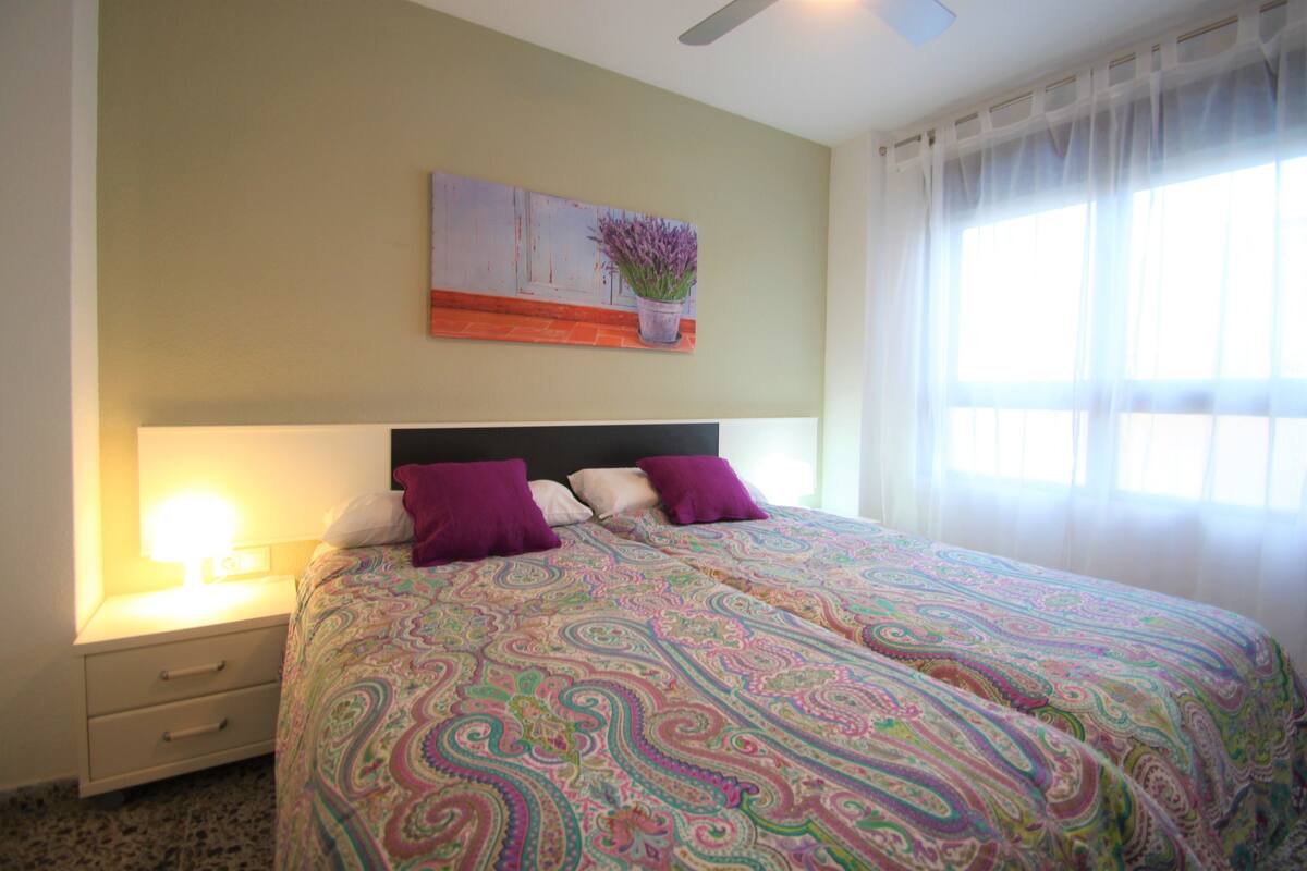 Une chambre à coucher avec un lit, une table de chevet, une lampe, un tableau et une fenêtre avec des rideaux.