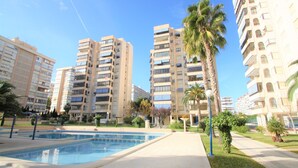 A pool area with a clear sky, surrounded by tall residential buildings and palm trees. - Apartamento CasaTuris Portobello (Alicante)