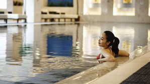 Mulher de fato de banho vermelho sentada na borda de uma piscina coberta com chão de azulejos e parede azul ao fundo.
