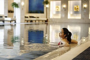 Una mujer con traje de baño rojo sentada al borde de una piscina interior, con suelo de baldosas y una pared azul de fondo.
