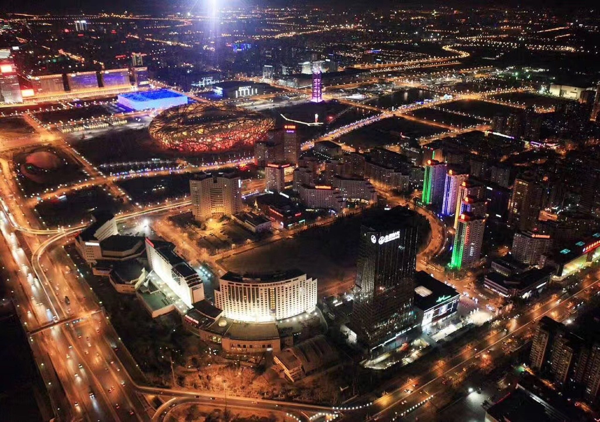 An aerial view of a cityscape at night with illuminated buildings, roads, and a stadium.