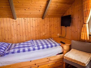 A wooden cabin room with a bed, a small table, a chair, and a television mounted on the wall.