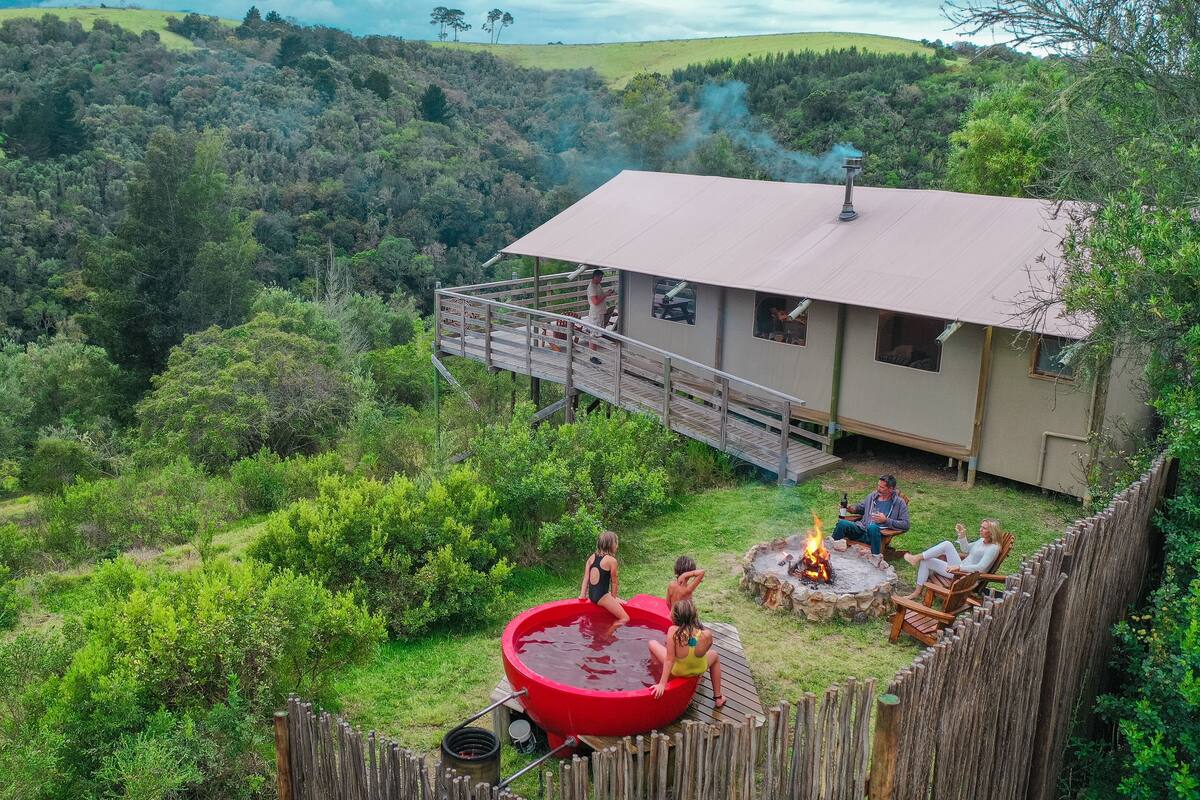 A house with a deck, a fire pit, and a red hot tub in a lush, green landscape.