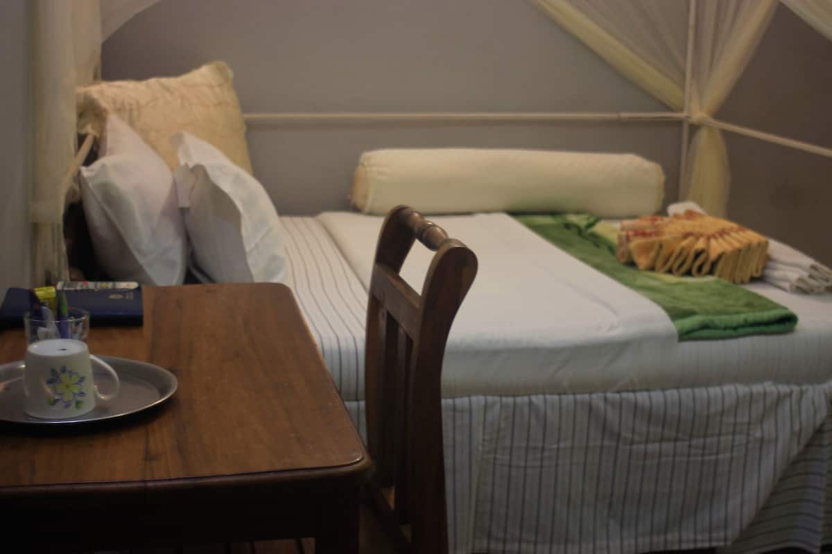 Quarto de hotel com uma cama, uma cadeira de madeira, uma mesa com bandeja e uma mesinha de cabeceira com uma xícara e um livro.