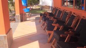 Une rangée de chaises en bois, dotées de coussins foncés, est disposée dans un espace extérieur abrité, dont le sol est recouvert de carreaux.