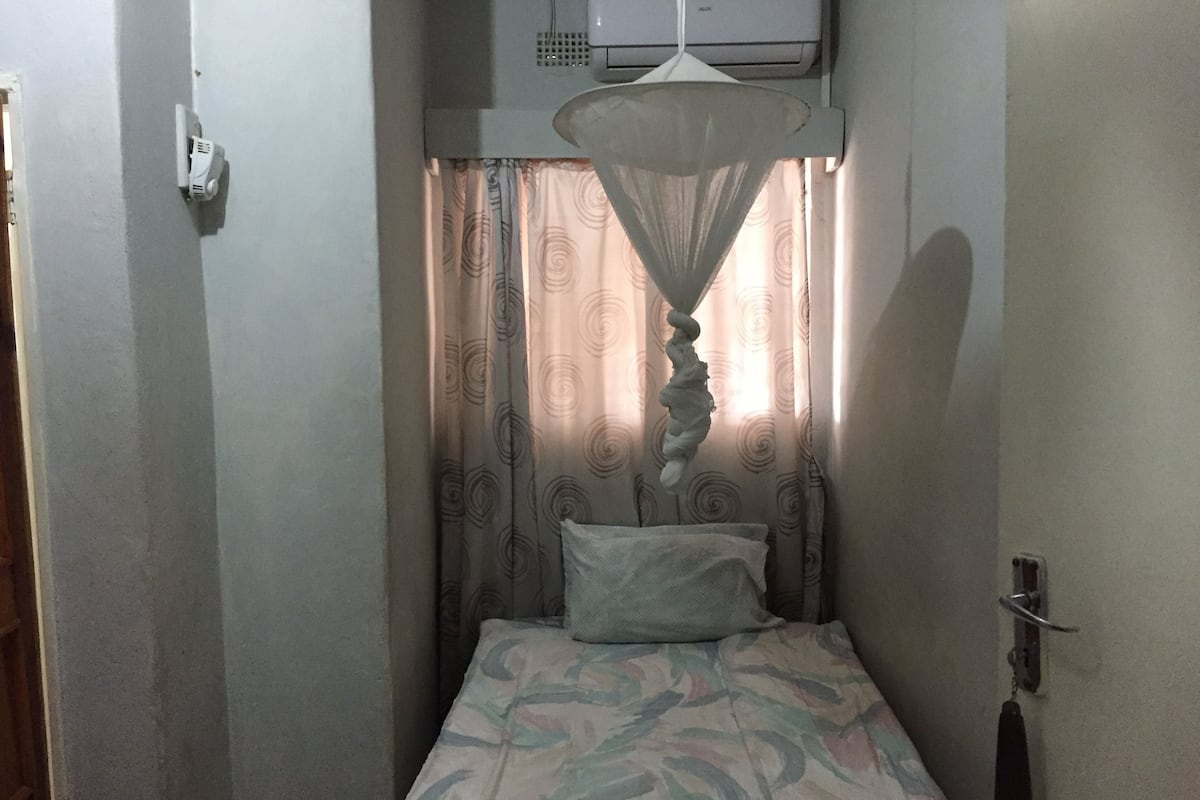 Um quarto pequeno e estreito com uma cama de solteiro, luminária pendente e janela com cortinas estampadas.